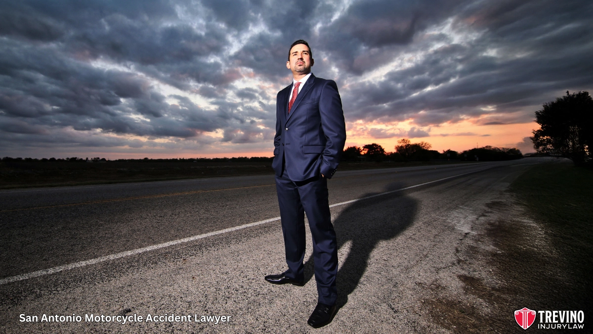 San Antonio Motorcycle Accident Lawyer, Candelario (J.R.) Trevino, wearing a blue suit stands confidently on the side of an empty rural road at sunset, with dramatic clouds overhead. The sun peeks through the horizon. Text reads “San Antonio Motorcycle Accident Lawyer” and includes the Trevino Injury Law logo.
