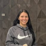 Trevino Injury Law Reviews. A woman with long dark hair smiles at the camera in a gray hoodie. The stylish background features a dark, geometric-patterned wall—much like the sharp design of San Antonio Personal Injury Lawyer Trevino Injury Law.