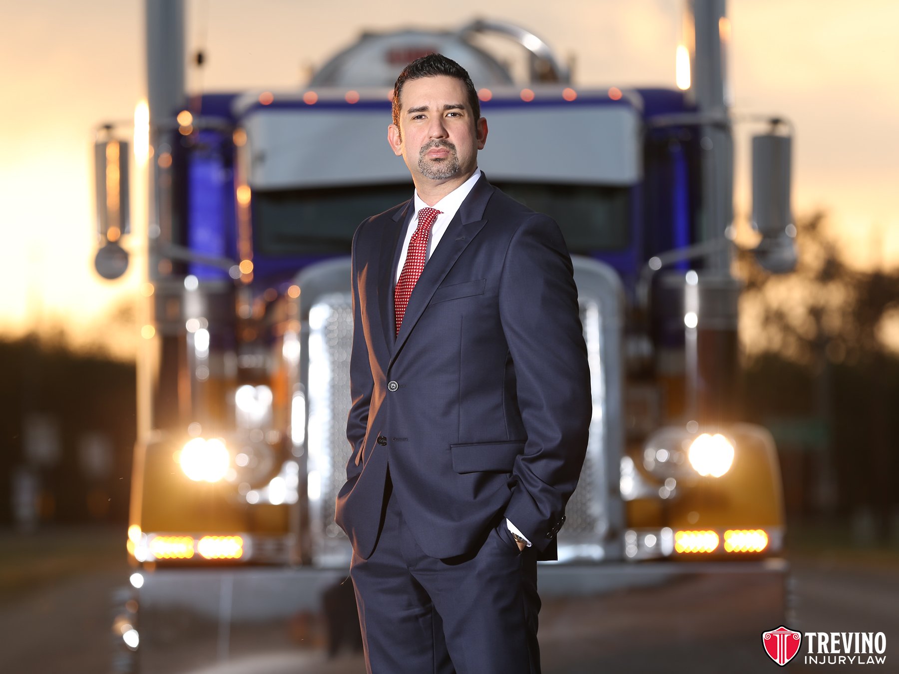 Best personal injury lawyer San Antonio. Attorney Candelario (J.R.) Trevino, owner of Trevino Injury Law in San Antonio stands confidently in a navy suit and red tie stands with hands in pockets before a large blue semi-truck at sunset. The truck's headlights are on. Trevino Injury Law, a San Antonio personal injury lawyer, logo appears in the lower right corner.