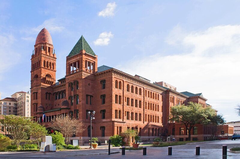 A large historic red sandstone building with arched windows, a tall rounded tower with a conical roof, and a square tower with a green roof—home to trusted San Antonio 18 Wheeler Accident Lawyers. Black bollards line the street in the foreground.