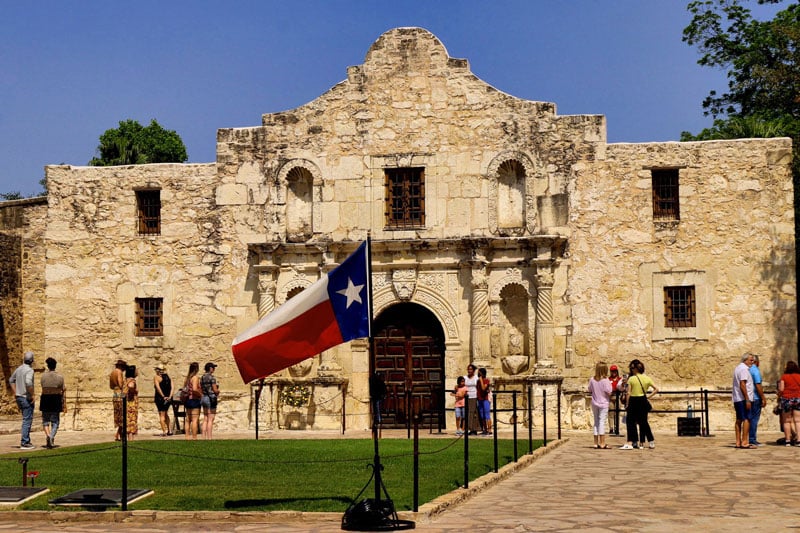 The image shows the historic Alamo mission in San Antonio, Texas, with visitors gathered outside. A prominent Texas state flag stands in the foreground—a familiar sight to locals, including San Antonio 18 Wheeler Accident Lawyer and Semi-Truck Attorneys.